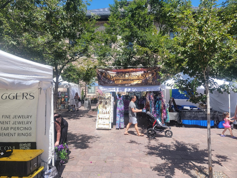Outdoor market scene with white vendor tents, trees, and a central stall displaying colorful jewelry. A person pushes a stroller through the market, with other shoppers and vendors visible in the background.