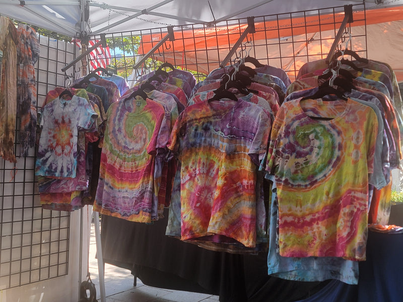 A display of colorful tie-dye t-shirts hanging on black wire racks at an outdoor market under a white canopy tent.