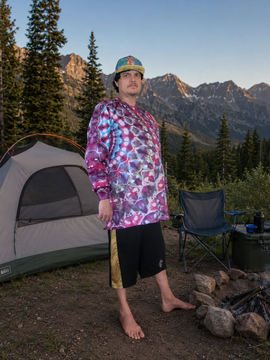 A man stands barefoot at a mountain campsite wearing a colorful purple, pink, and blue tie-dye long-sleeve shirt, black shorts, and a green cap. A gray tent, folding camp chair, and fire pit are visible in the background with pine trees and mountains under a blue sky.