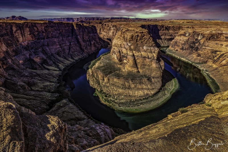 A high view of a river bending in a tight horseshoe around a sandstone mesa, with purple twilight clouds and a thin green shoreline.