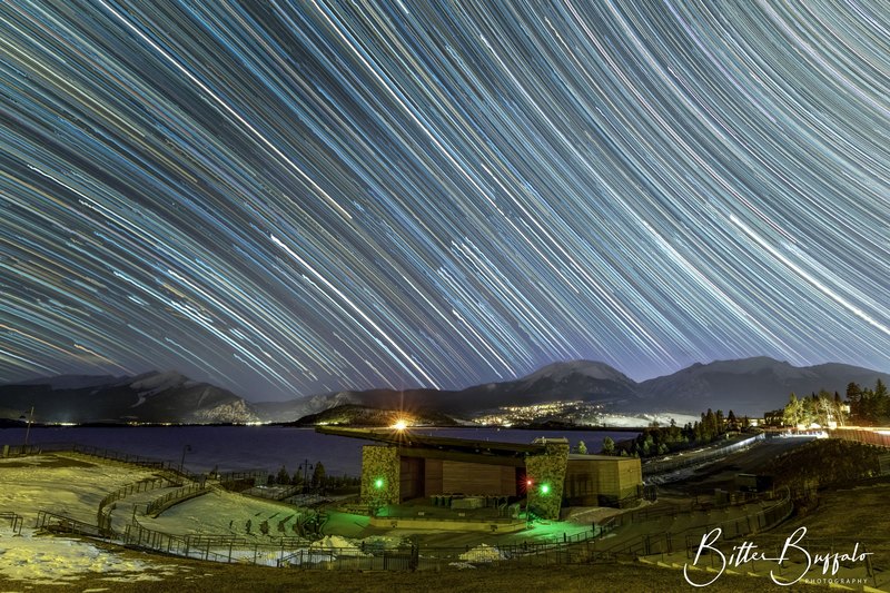 Long-exposure star trails streak across a night sky above snow-dusted mountains, a lake, and an outdoor amphitheater with green and red stage lights.