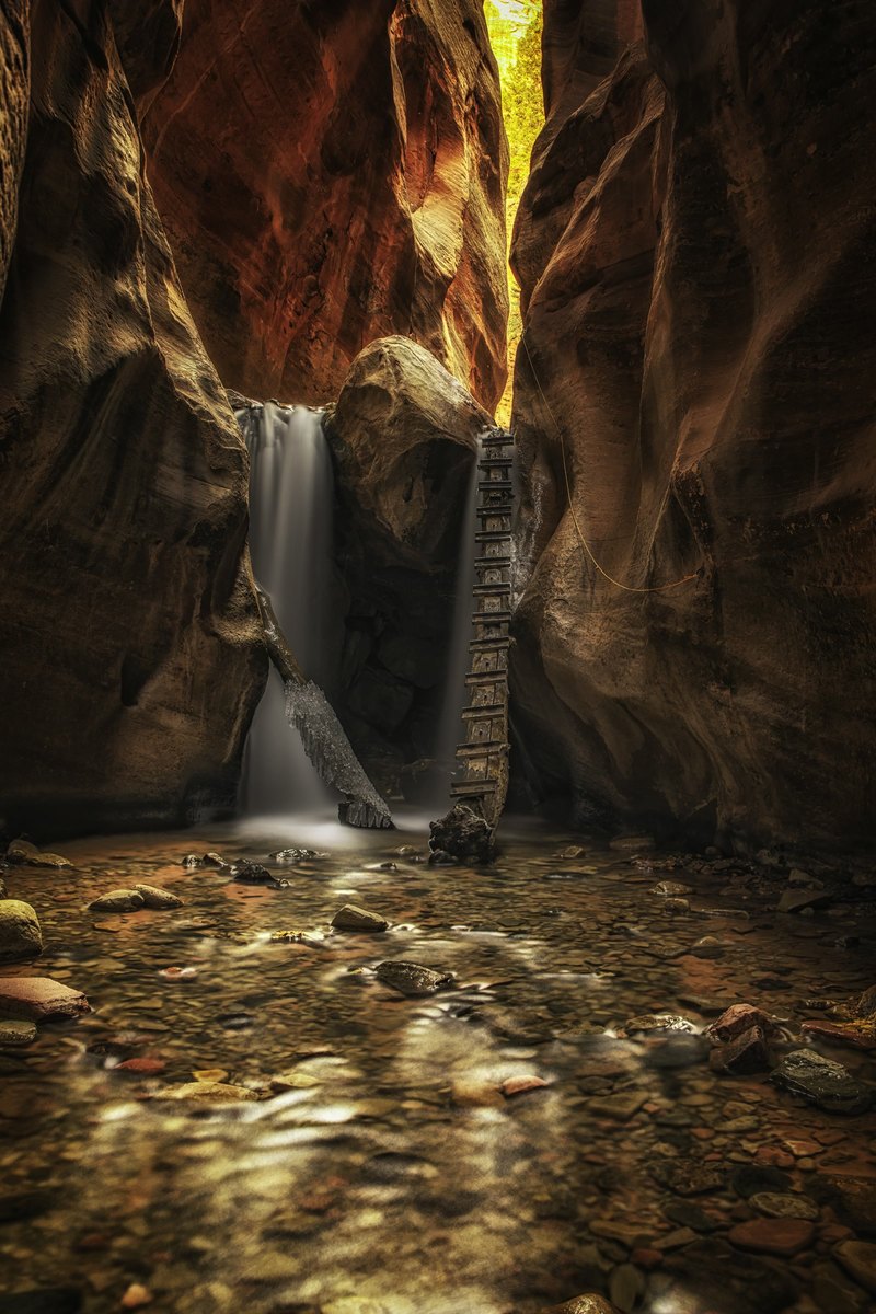 A vertical view inside a narrow sandstone canyon with a silky waterfall, a rustic wooden ladder, a large boulder, and a pebble-filled stream in the foreground.