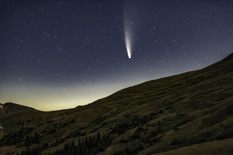A bright comet with a long pale tail hangs over a dark alpine hillside and distant mountain silhouettes, with a warm glow on the left horizon.