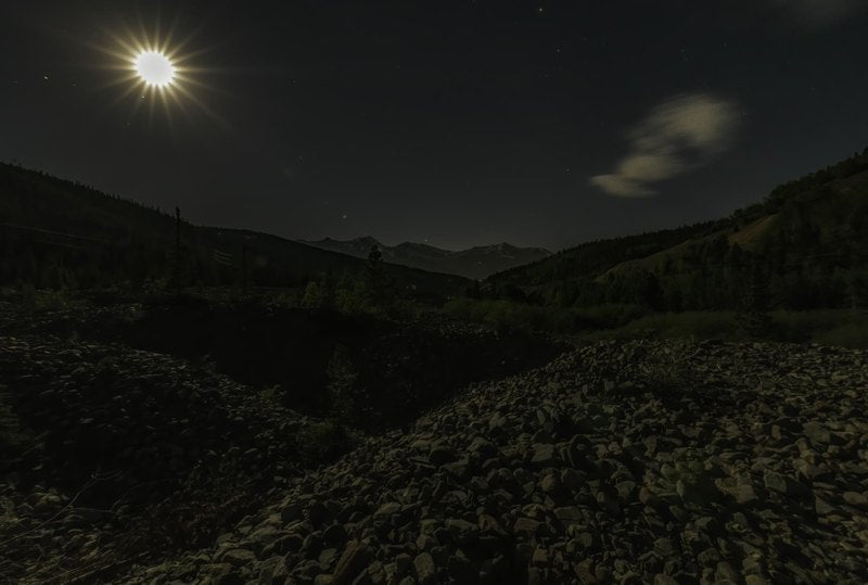 A bright starburst moon hangs over a rugged foreground of dark boulders, shadowed hills, and a distant jagged mountain range in a deep night sky.