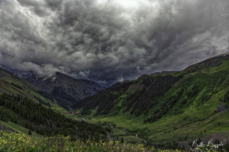 Heavy swirling storm clouds fill the sky above a lush green mountain valley with a thin winding stream and wildflowers along the bottom edge.