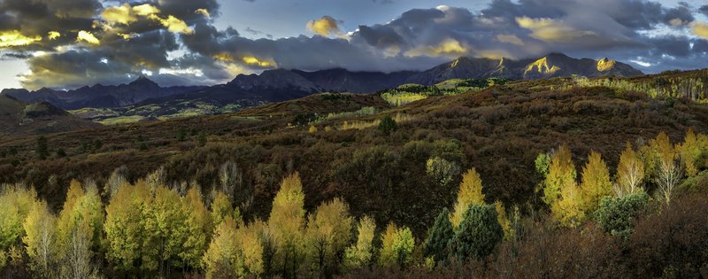 A wide panorama of yellow aspens in the foreground, dark rolling hills, and a jagged mountain range under sunlit storm clouds.