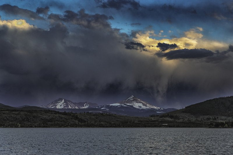 Snow-dusted triangular peaks rise above a dark forested ridge and charcoal lake, with storm clouds, a shaft of rain, and a golden break of light at right.