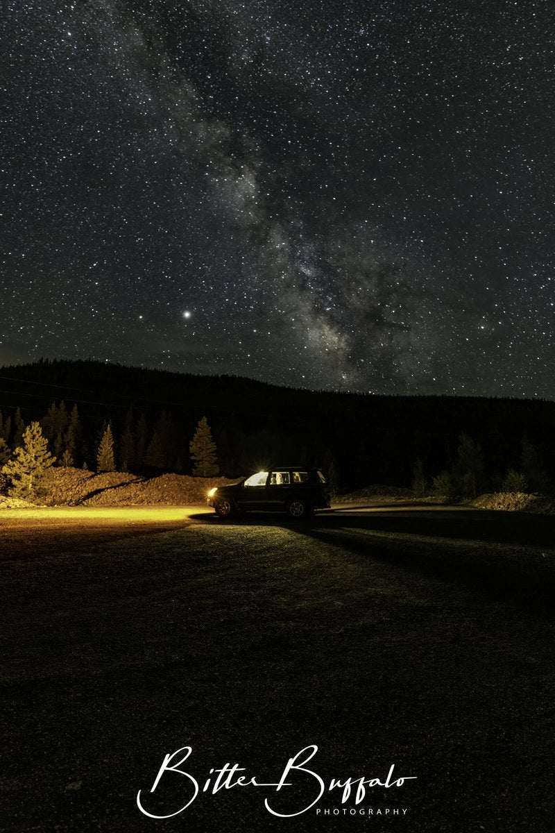 A vertical night photograph of the Milky Way above a dark SUV on a gravel clearing, with warm vehicle lights on the ground and a silhouetted treeline.