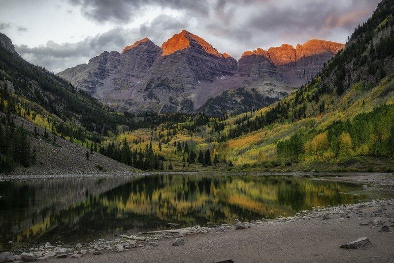 Twin reddish mountain peaks and yellow aspens are reflected in a still alpine lake at dawn, with driftwood on the shore and dramatic clouds above sunlit ridges.