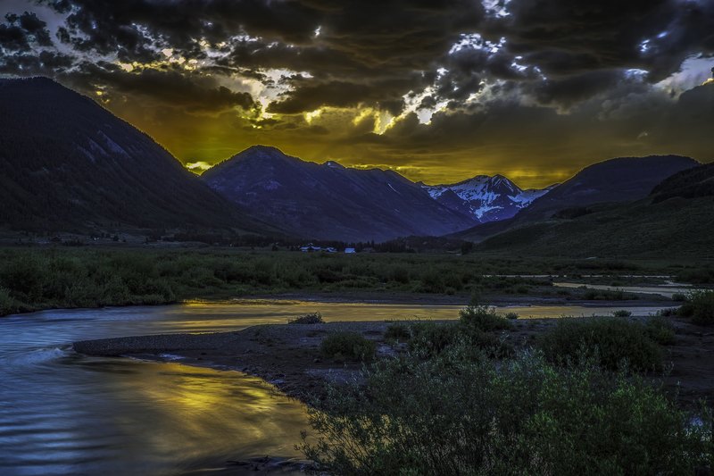 A winding river reflects golden sunset light in a green mountain valley, with dark storm clouds, a snow-patched peak, and small white buildings in the distance.