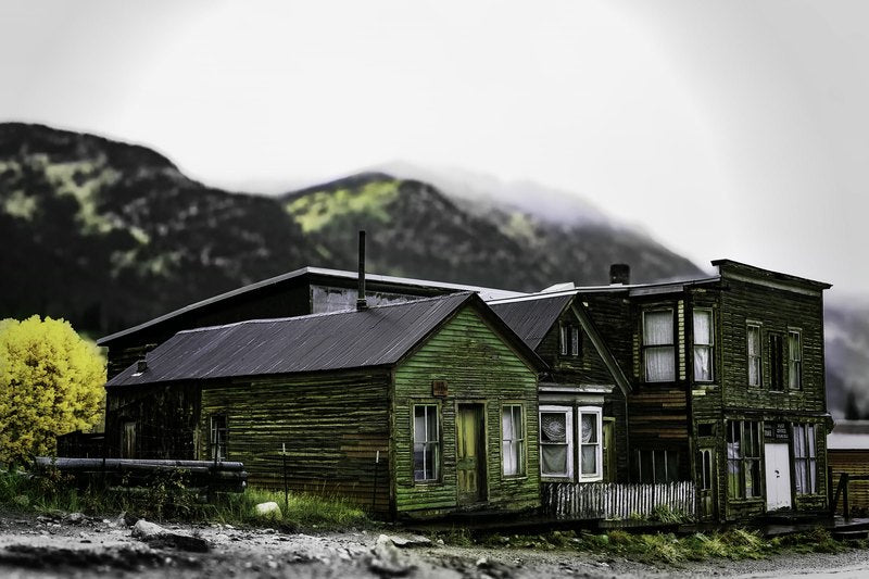 A faded green gabled wooden building stands beside a taller dark wooden storefront at the base of misty mountains, with gravel in the foreground.