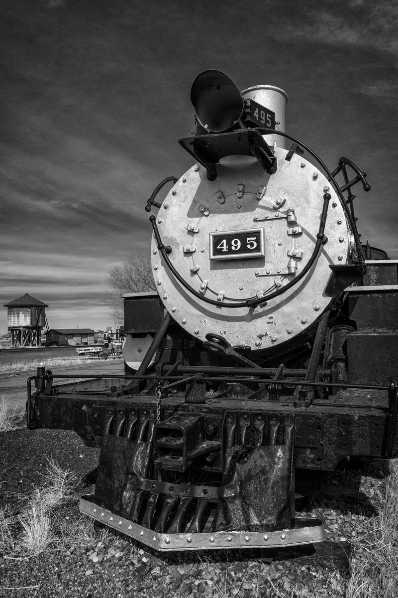 A black-and-white head-on view of steam locomotive 495, showing the bolted boiler plate, headlight, and cowcatcher, with a wooden water tower in the distance.