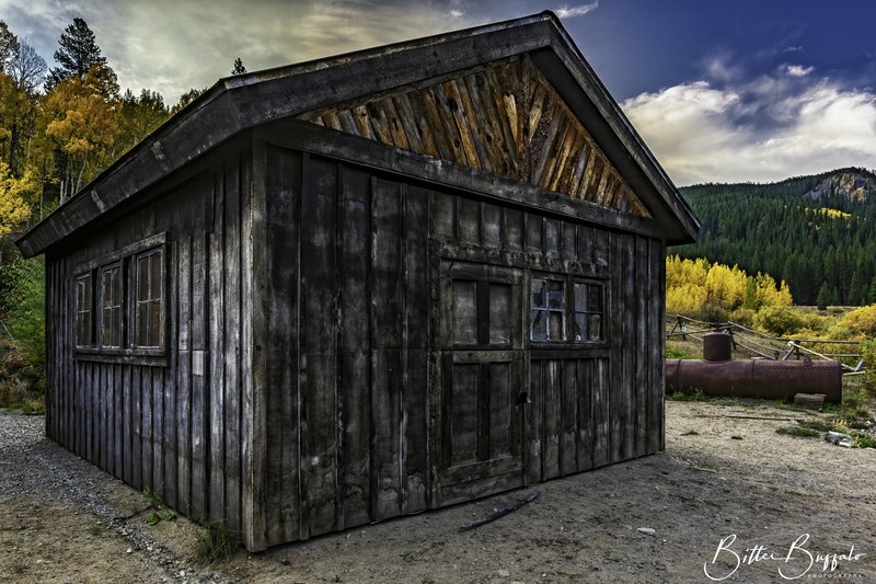 A dark weathered wooden outbuilding with a decorative sunburst gable stands beside a rusted tank and log fence, with golden aspens and a mountain ridge behind.