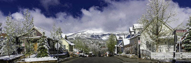 A wide view of a snow-dusted historic street with a French St and National Historic District sign, Victorian houses, and a snow-capped mountain under a blue sky.