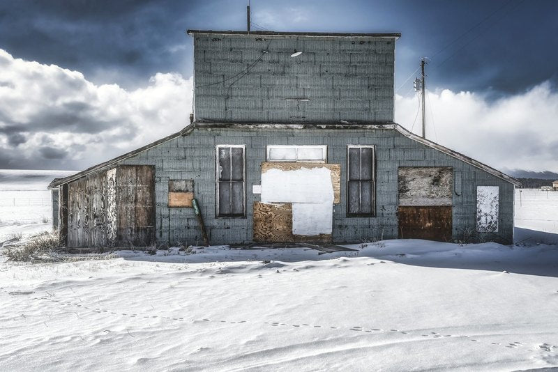 An abandoned two-story weathered teal building with boarded windows stands in a snow field under a dramatic storm sky, with animal tracks in the snow.