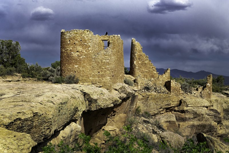 Crumbling tan stone ruins with a circular tower sit on a rocky desert ledge under dark storm clouds, with a black bird perched on the highest stones.