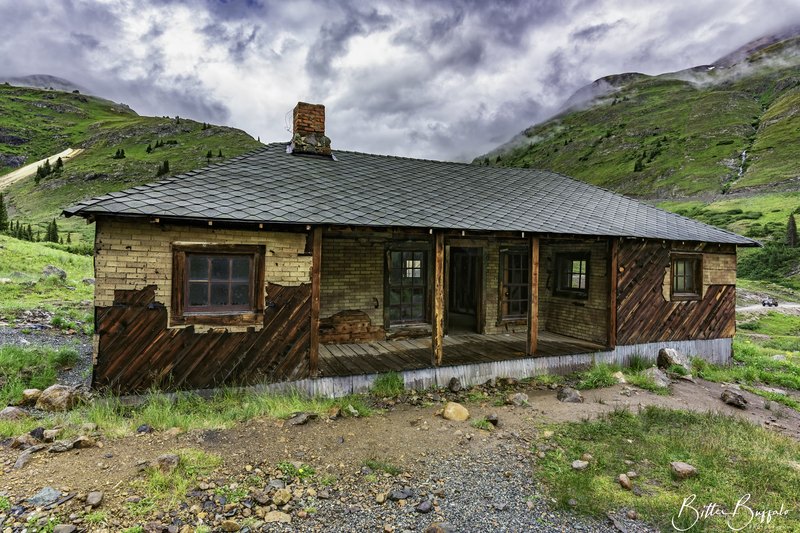 A small dilapidated cabin of brick, diagonal wood, and corrugated metal sits on rocky ground in a green mountain valley under heavy clouds.