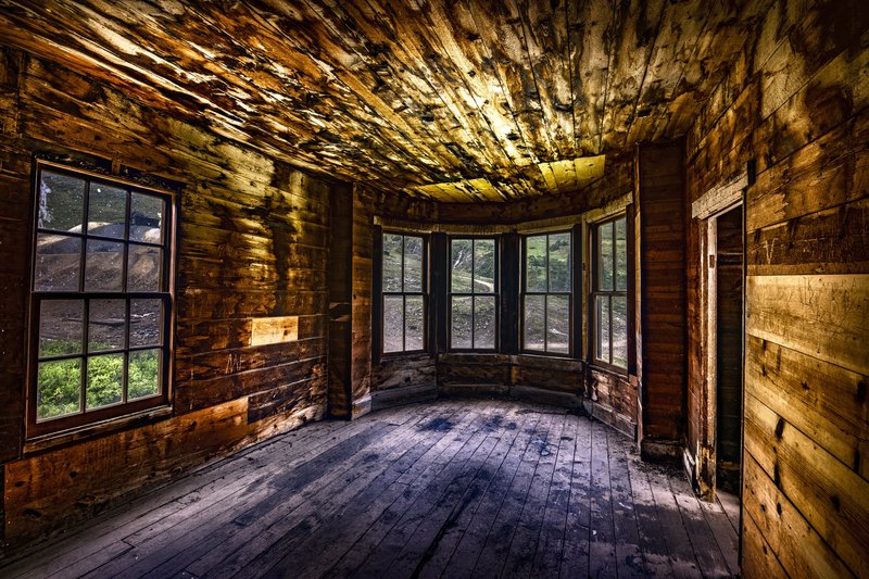 The empty wooden interior of an abandoned building, with golden light on the ceiling, cool blue shadows on the floor, and mountain greenery visible through old windows.