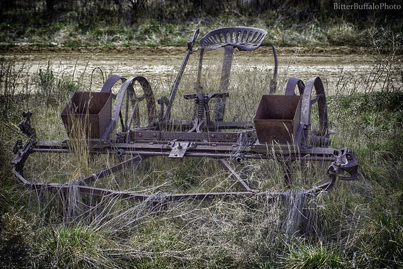 A heavily rusted antique two-row planter with a metal seat and seed hoppers sits in tall dry grass, with a blurred dirt path and trees behind it.