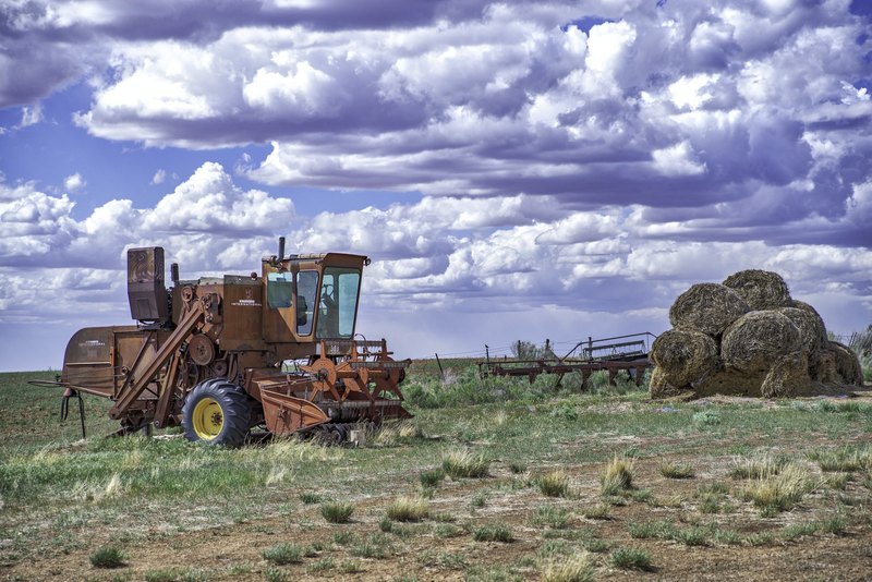 A rusty International Harvester combine with a yellow wheel rim sits in a dry field beside stacked round hay bales under a large cloudy sky.