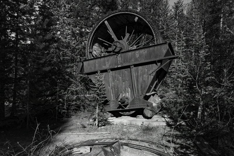 A black-and-white photograph of a large riveted metal hopper with a spoked wheel on top, sitting on logs in a dense pine forest with rails in the foreground.