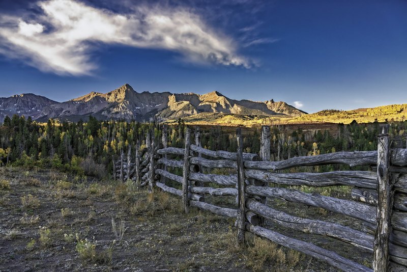 A weathered buck-and-rail log fence runs diagonally toward a valley of yellow aspens and dark pines, with sunlit jagged peaks under a deep blue sky.