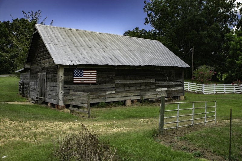 A long silver-grey wooden barn with a corrugated metal roof and an American flag on its side sits in a grassy field beside a white rail fence.